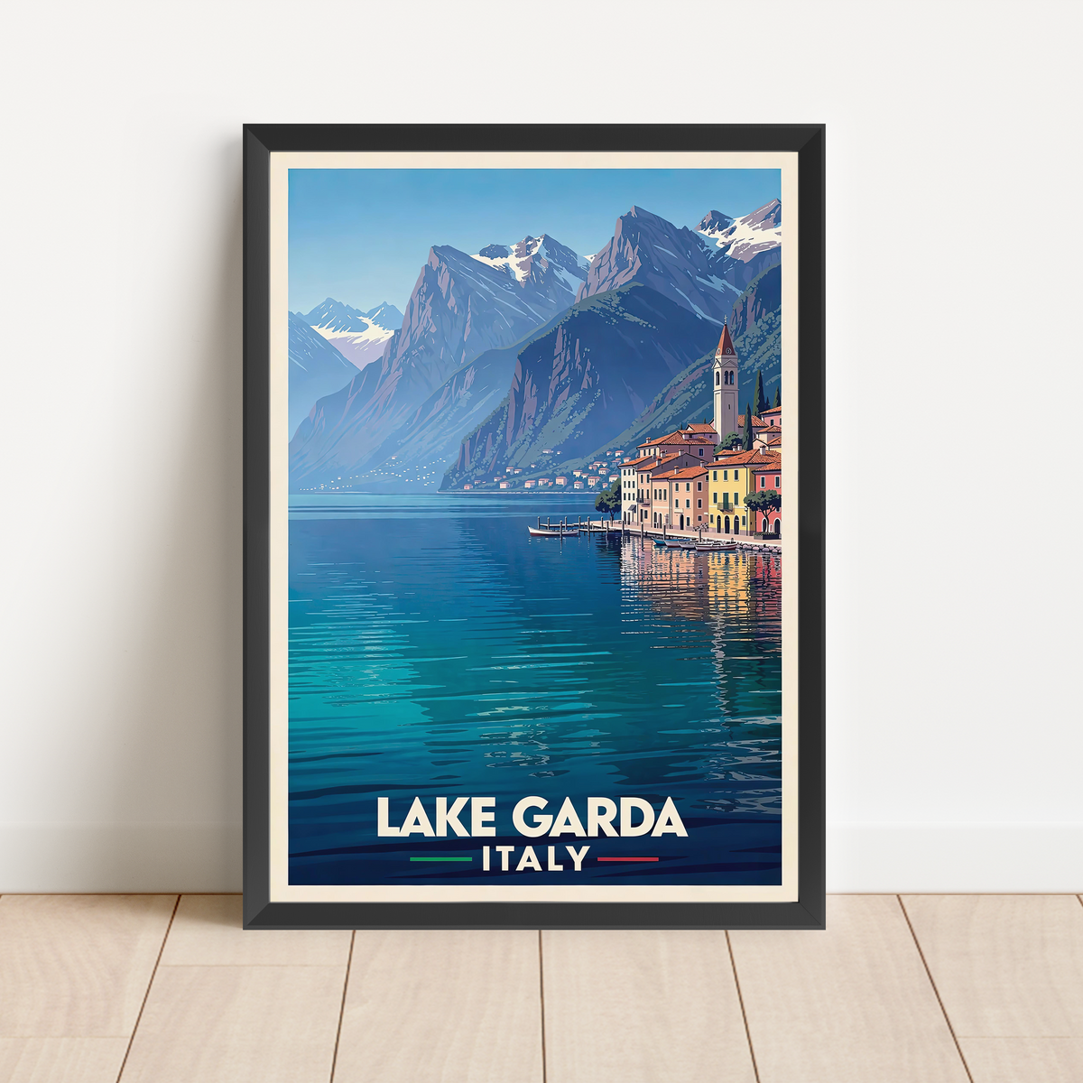 A framed travel poster of Lake Garda, Italy, leaning against a white wall on a light wood floor. The poster features a vintage-style illustration of the blue lake, mountains, and colorful buildings, with 'LAKE GARDA ITALY' printed at the bottom.
