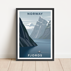 A framed travel poster of Norway's fjords, leaning against a white wall on a light wood floor. The poster features a stylized illustration of blue water, steep gray mountains, and snow-capped peaks, with the words 'NORWAY' at the top and 'FJORDS' at the bottom.